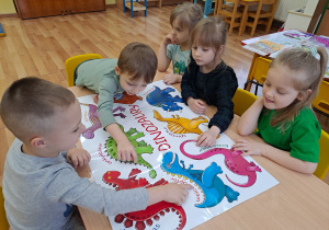 Trzy dziewczynki i dwóch chłopców oglądają kolorowy plakat z dinozaurami.